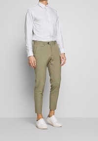 Lichtgrijs en wit gestreepte button-up shirt, op maat gemaakte beige broeken en witte sneakers. Opvallende kenmerken zijn de smalle pasvorm en omgeslagen mouwen.