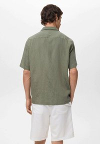 Olive green short-sleeve shirt with vertical white stripes, featuring a relaxed fit and a folded collar. Worn with white shorts.