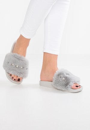 Pieds portant un pantalon blanc et des sandales slides grises en fourrure ornées de petites perles sur un sol blanc.