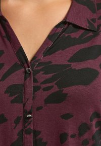 Primo piano di una camicia viola con un motivo astratto nero, dotata di un colletto e due bottoni neri lucidi lungo la pattina.