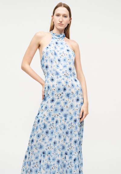 Γυναίκα με ίσια μαλλιά που φοράει αμάνικο φόρεμα με λαιμόκοψη halter και μπλε ανθισμένο σχέδιο σε λευκό φόντο.
