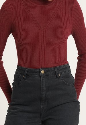 Personne portant un pull cintré côtelé à manches longues bordeaux rentré dans un jean noir taille haute avec fermeture par bouton.