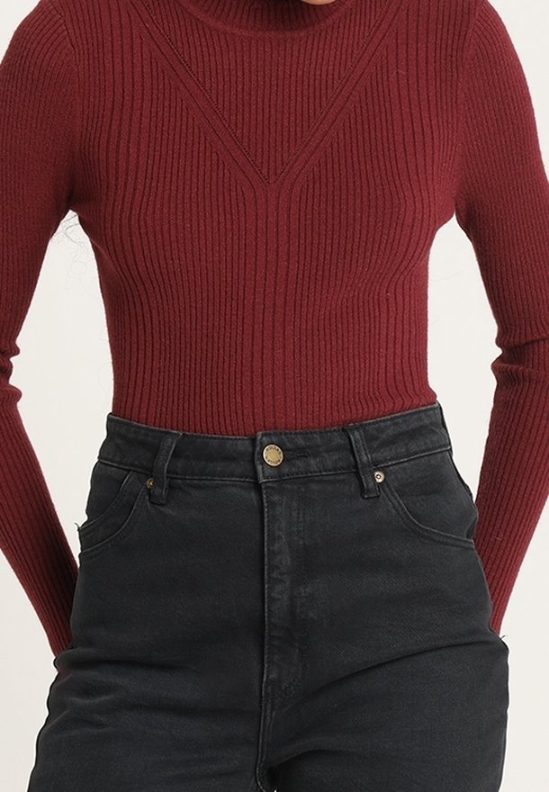 Personne portant un pull cintré côtelé à manches longues bordeaux rentré dans un jean noir taille haute avec fermeture par bouton.