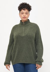 Suéter de punto verde bosque con cuello alto y cierre de media cremallera. Textura acanalada, corte relajado y mangas largas. Combinado con jeans azules.