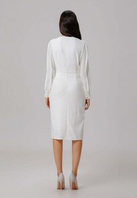 Robe blanche à longueur genou avec des manches bouffantes, une silhouette ajustée et une fermeture éclair dans le dos. Associée à des chaussures à talons hauts gris clair.