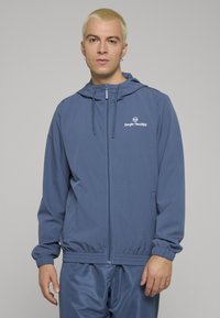 Kék sportdzseki cipzáros elülső résszel és kapucnival. Rugalmas mandzsettákkal és szegéllyel rendelkezik, valamint fehér logó található a mellrészen. Könnyű anyagból készült.