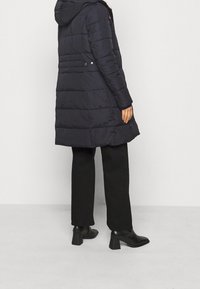 Marinblå pufferjacka med huva, försedd med horisontell quiltning, knäppningar på sidorna och en något utsvängd nederkant. Matchad med svarta byxor och stövlar med klack.