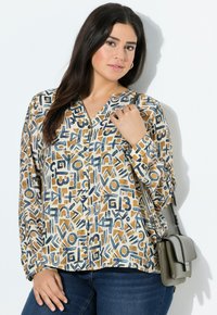 Frau trägt ein gemustertes, knopfbares Hemd mit blauen und beigen geometrischen Formen, dunkelblaue Jeans und eine graue Umhängetasche.