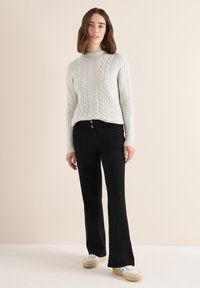 Jasnoszary sweter w splot warkoczowy z wysokim kołnierzem, zestawiony z czarnymi spodniami o rozszerzanych nogawkach. Modelka nosi kremowe sneakersy z niebieskimi akcentami.