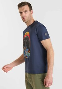 Hombre vistiendo una camiseta azul marino con un gráfico de calavera colorido, mariposas y flores, combinado con pantalones verdes oliva, de pie de lado.
