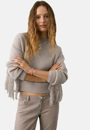 Gray ribbed sweater with fringe details at the sleeves, paired with light gray pants. The sweater has a cropped design and textured fabric.