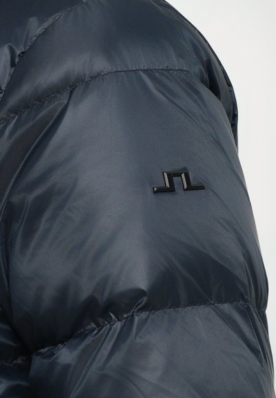 Dunkelblau gesteppte Jacke mit glänzender Textur, ausgestattet mit einem schwarzen Logoakzent am linken Ärmel und genähten Details entlang der Nähte.