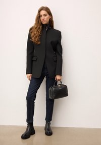 Črn tailored blazer z visokim ovratnikom, v kombinaciji s temnimi denim hlačami in masivnimi črnimi gležnjarmi. Drži majhno črno torbico.