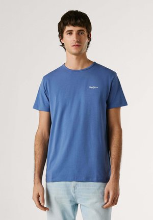 Jonge man staand, gekleed in een blauw T-shirt met korte mouwen en lichtblauwe jeans, voor een effen lichte achtergrond.