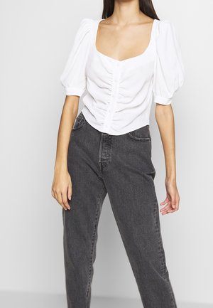 Blusa bianca con maniche a sbuffo, scollatura quadrata, bottoni frontali e tessuto strutturato, abbinata a jeans grigi scuri a vita alta.