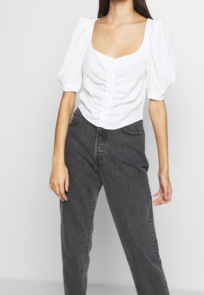 Bílá blůza s nadýchanými rukávy a čtvercovým výstřihem, s předním zapínáním na knoflíky a texturovanou látkou, kombinovaná s tmavě šedými džíny s vysokým pasem.