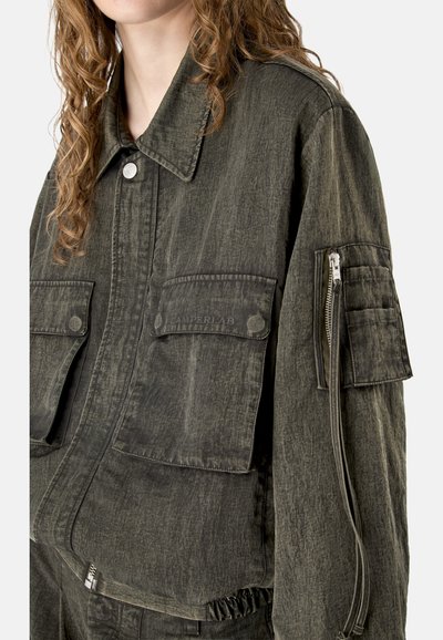 Mujer con chaqueta texturizada verde oscuro con grandes bolsillos delanteros con solapa y bolsillo con cremallera en la manga izquierda, con el cabello rizado visible.