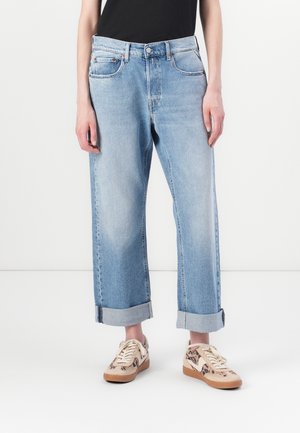 Světle modré ohrnuté džíny kombinované s teniskami se zvířecím potiskem a černým topem, zobrazené od pasu dolů na bílém pozadí.