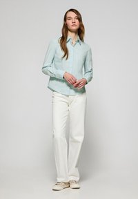 Camisa de botón azul clara con cuello, mangas largas y un corte relajado, combinada con pantalones blancos de pierna recta y zapatillas beige.