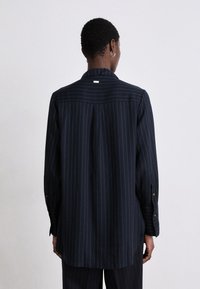 Navy striped shirt with a collar, long sleeves, and buttoned cuffs. It features a straight cut and a prominent back pleat.