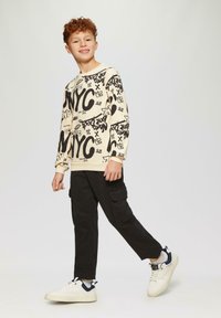 Beżowa bluza z nadrukiem czarnego tekstu NYC, w połączeniu z czarnymi spodniami cargo. Model nosi białe sneakersy z granatowymi akcentami.