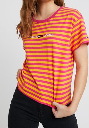 Femme portant un t-shirt Tommy Jeans à manches courtes rayé rose et orange, associé à un pantalon noir, tenant l'ourlet du t-shirt.