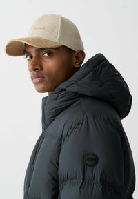 Črna napihnjena jakna s kapuco, s šivanimi paneli in okroglo črno oznako. Sivi kordurojski klobuk z ukrivljenim robom in vezenjem.