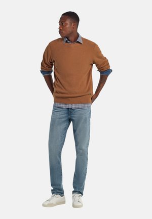 Uomo con un maglione marrone sopra una camicia a quadri blu, jeans azzurri e sneakers bianche, in piedi con le mani dietro la schiena.