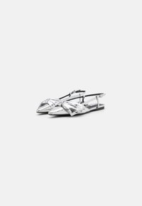 Scarpe piatte metalliche argento con punta affusolata e design frontale annodato. Presentano una cinghia alla caviglia e una suola piatta. Texture liscia.