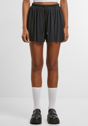 Vrouw die zwarte geplooide korte broek met trekkoord, witte geribbelde sokken en zwarte loafers met plateauzool draagt, staand voor een effen achtergrond.