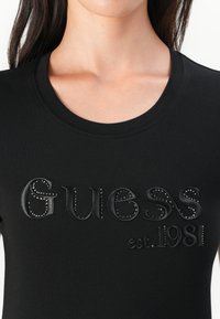 Black cotton T-shirt featuring textured black logo "Guess" with studs, round neckline, and short sleeves, highlighting a fitted design.