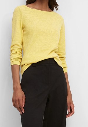 Kobieta ubrana w żółtą bluzkę z długim rękawem, wsuniętą w czarne spodnie z wysokim stanem, stojąca na tle jednolitej białej ściany.