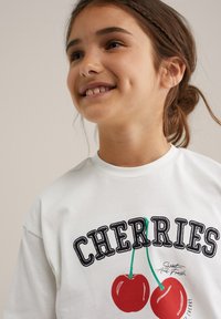 Balta medvilninė marškinėlių su grafika, ant kurios pavaizduotos dvi raudonos vyšnios su žaliomis kotelėmis, o viršuje yra tekstas "VYŠNIOS" paryškintais juodaisiais rašmenimis.