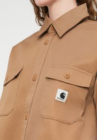 Καφέ πουκάμισο με κουμπιά από ανθεκτικό ύφασμα, διαθέτει δύο τσέπες στο στήθος, εμφανείς ραφές και τετράγωνο λογότυπο στην αριστερή τσέπη.