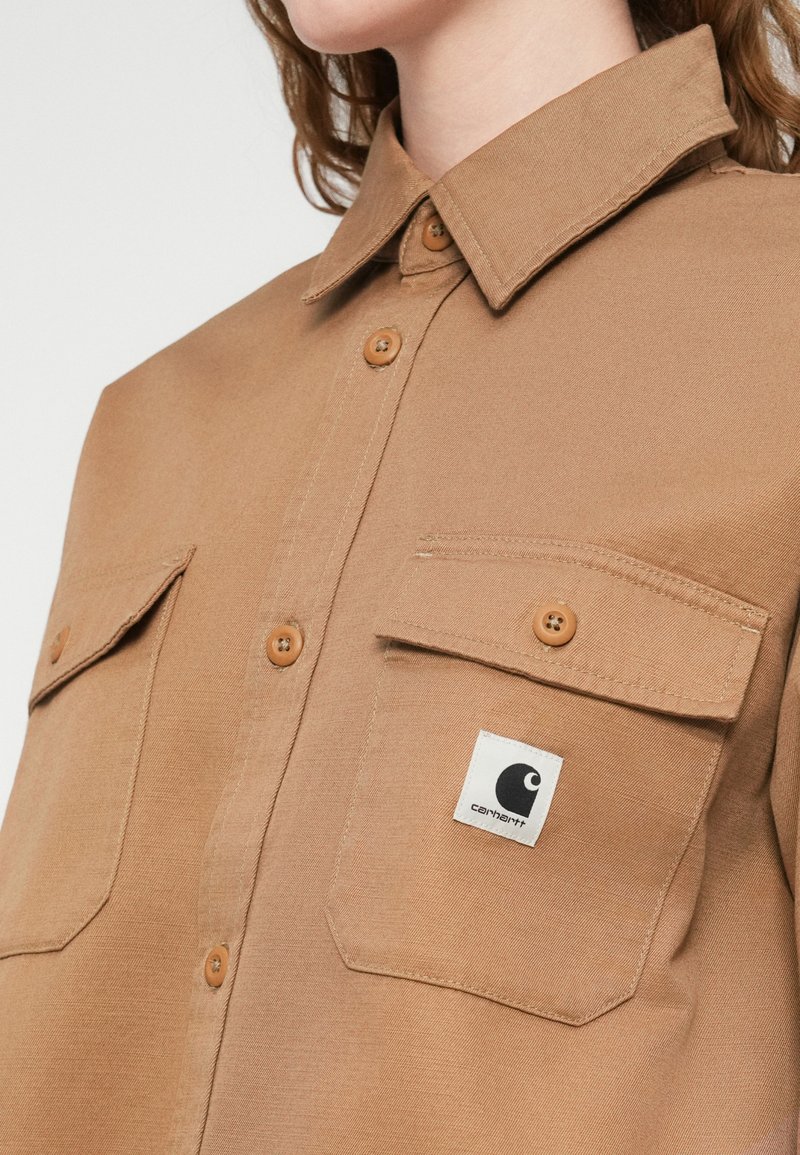 Braunes Hemd zum Knöpfen aus robustem Stoff, mit zwei Brusttaschen, auffälligen Nähten und einem quadratischen Logo-Patch auf der linken Tasche.