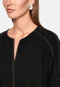 Schwarzes Oberteil mit V-Ausschnitt und kontrastierenden weißen Nähten. Verfügt über einen glatten, leichten Stoff und wird mit einem glänzenden silbernen Ohrring getragen.