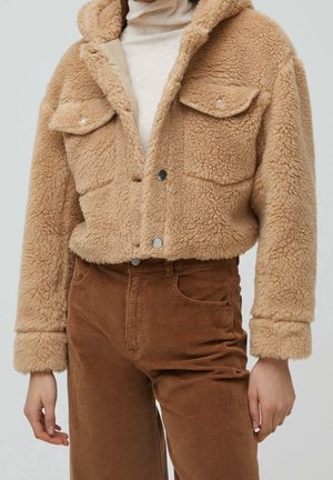 Personne portant une veste sherpa beige avec des boutons-pression sur un col roulé blanc et un pantalon en velours côtelé marron.