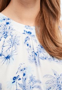 Witte blouse met een blauwe tropische print van palmbomen en dieren. De stof heeft een gestructureerd patroon, aangevuld met een geplooide halslijn.