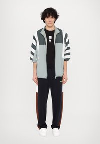 Lyseblå og hvid stribet zip-op jakke over en sort t-shirt, parret med mørke bukser med brune og hvide detaljer og hvide sneakers.