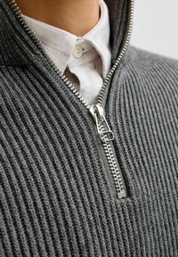 Tuvumā attēlots pelēks ribots adījuma jaka ar rāvējslēdzēju, kas uzvilkta virs baltas svītrainas krekla apkakles.