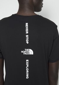 Hombre con una camiseta negra con texto vertical en blanco que dice "Nunca dejes de explorar" y el logo de The North Face en la espalda.