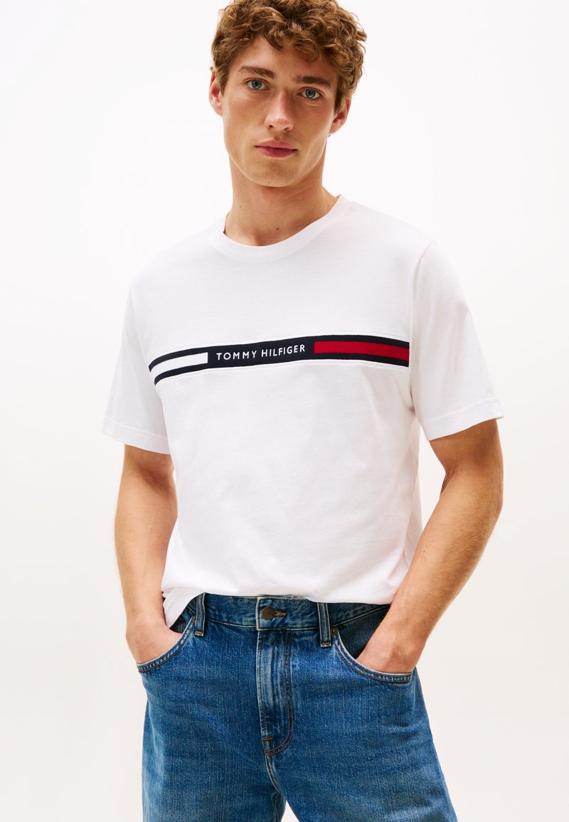 Fiatal férfi göndör hajjal, fehér Tommy Hilfiger pólót visel, amelyen logós csík fut a mellkasán, kék farmert és zsebre tett kezeket.