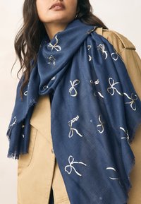 Sciarpa blu navy con trama testurizzata, decorata con motivi a fiocco dorati metallizzati. I bordi sono sfrangiati per una finitura morbida. Indossata drappeggiata intorno al collo.