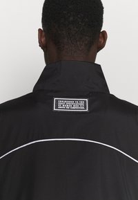 Chaqueta negra con un cuello alto, con ribetes blancos y una etiqueta en la parte posterior que muestra texto. Textura de tela suave y ligera.