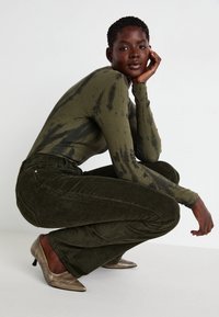 Maglia a maniche lunghe verde oliva con un motivo tie-dye abbinata a pantaloni a zampa d'elefante in velluto a coste verde scuro e scarpe con tacco metalliche.