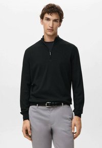 Musta vetoketjulla varustettu poolopaita, valmistettu pehmeästä kankaasta, jossa pitkät hihat ja rento istuvuus, yhdistetty vaaleanharmaisiin housuihin ja mustaan vyöhön.