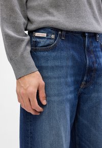 Jeans in denim blu con lavaggio medio, caratterizzati da cuciture gialle e un'etichetta Calvin Klein sulla tasca posteriore. Accompagnati da un maglione grigio.