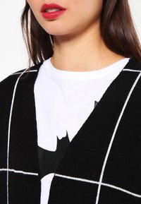 Svart cardigan med vitt rutmönster, lagt över en vit T-shirt med svart grafisk design. Slät textur, V-ringning.