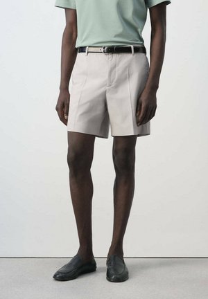 Hombre que lleva pantalones cortos plisados beige con cinturón negro, camisa verde y zapatos negros tipo slip-on, de pie sobre un suelo gris.