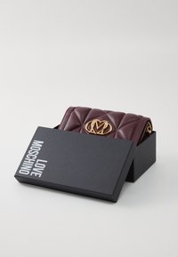 Burgundijas krāsas pārklāta āda somiņa melnā kastē. Iespējas ar zelta logo un ķēdes akcentiem. Kaste marķēta ar "Love Moschino."